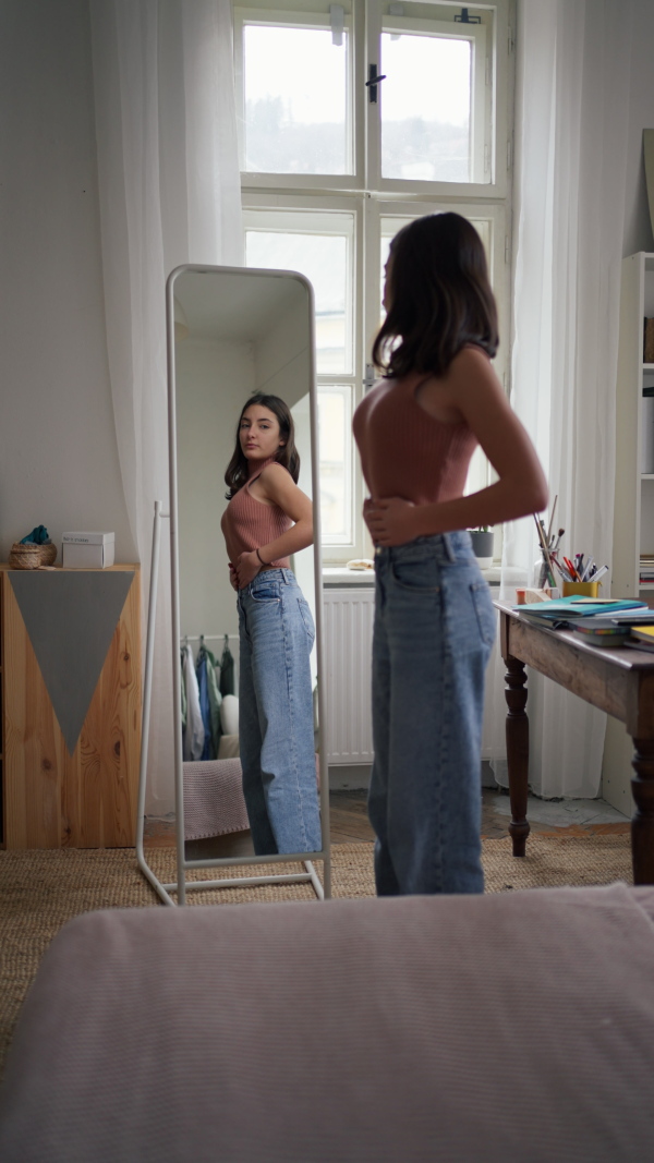 Vertical view of a young teenage girl looking in the mirror in the room, trying on clothes.