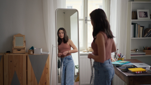 A young teenage girl looking in the mirror in the room, trying on clothes and measuring her waist.