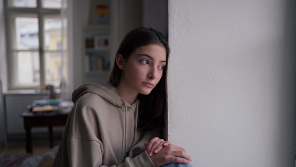 Unhappy teenage girl sitting in home and looking out of the window.