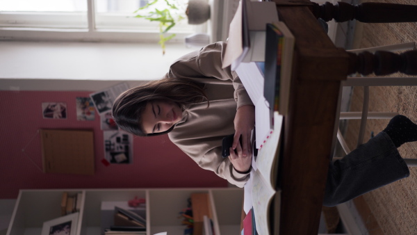 Vertical view of a young teenage girl studying in her room and scrolling smartphone.