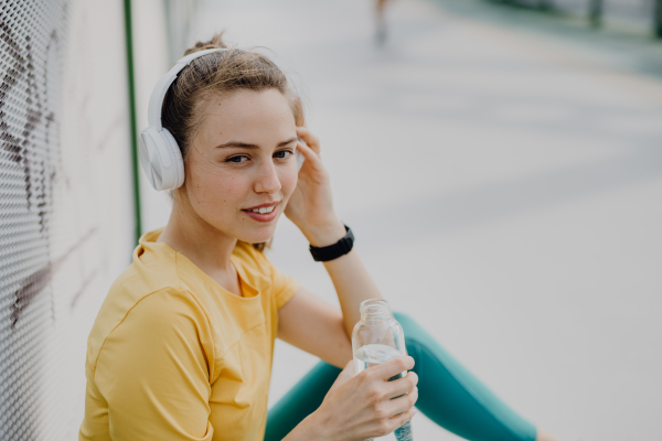 Young woman drinking water and listening the music trough headphones, during jogging in city, healthy lifestyle and sport concept.