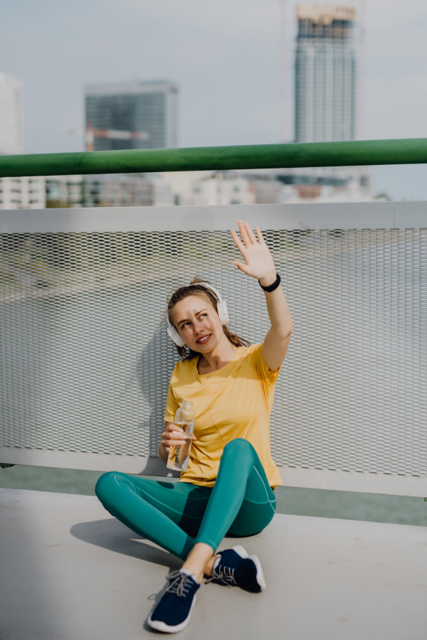 Young woman resting during city run, sitting and drinking water, listening music trough the headphones, healthy lifestyle and sport concept.