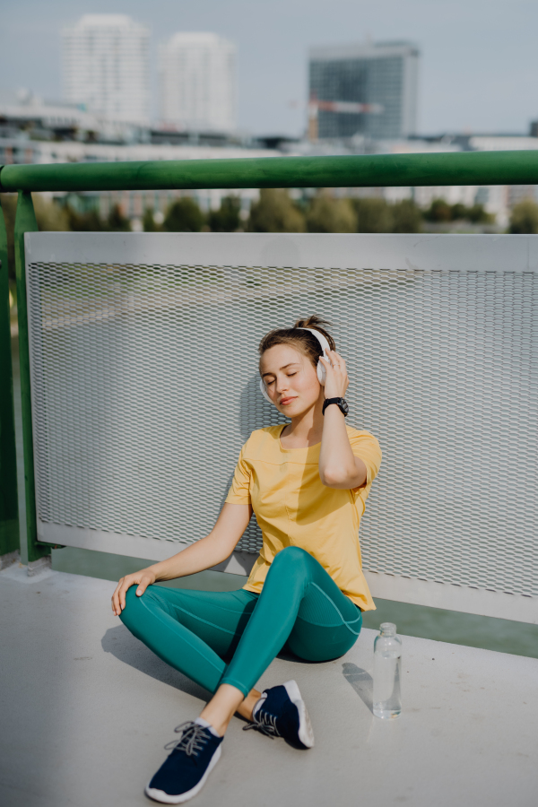 Young woman drinking water and listening the music trough headphones, during jogging in city, healthy lifestyle and sport concept.