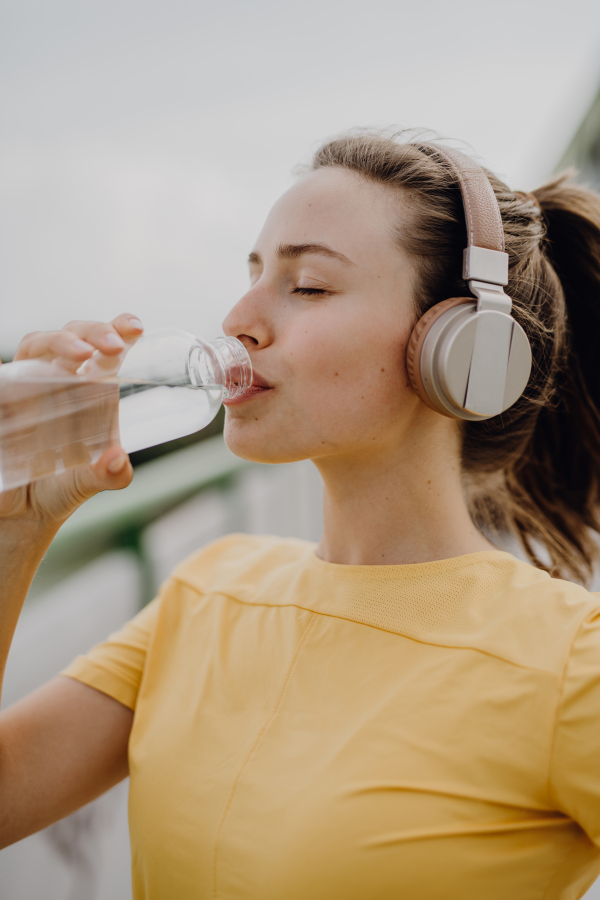 Young woman drinking water and listening the music trough headphones, during jogging in city, healthy lifestyle and sport concept.