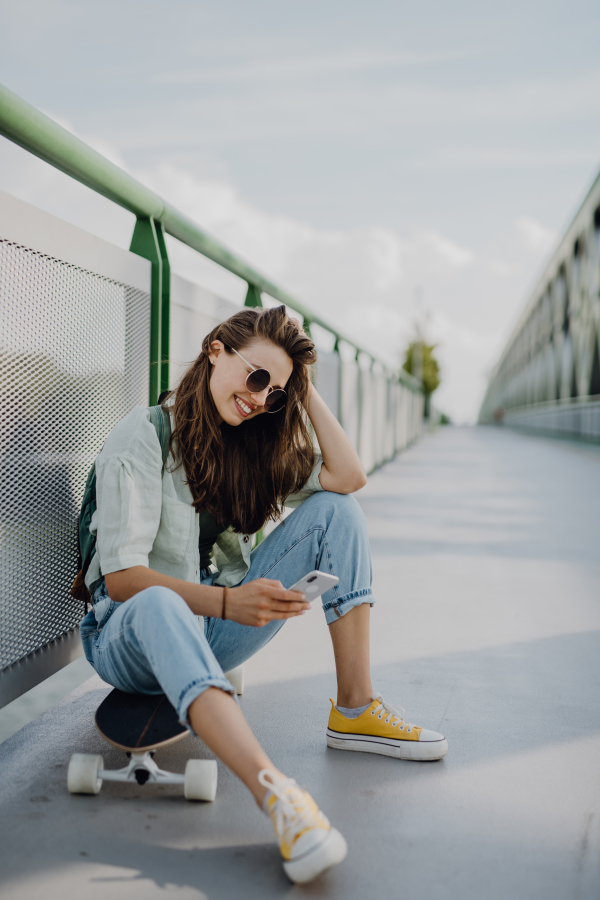 Happy young woman sitting on a skateboard and scrolling smartphone at city bridge. Youth culture and commuting concept.