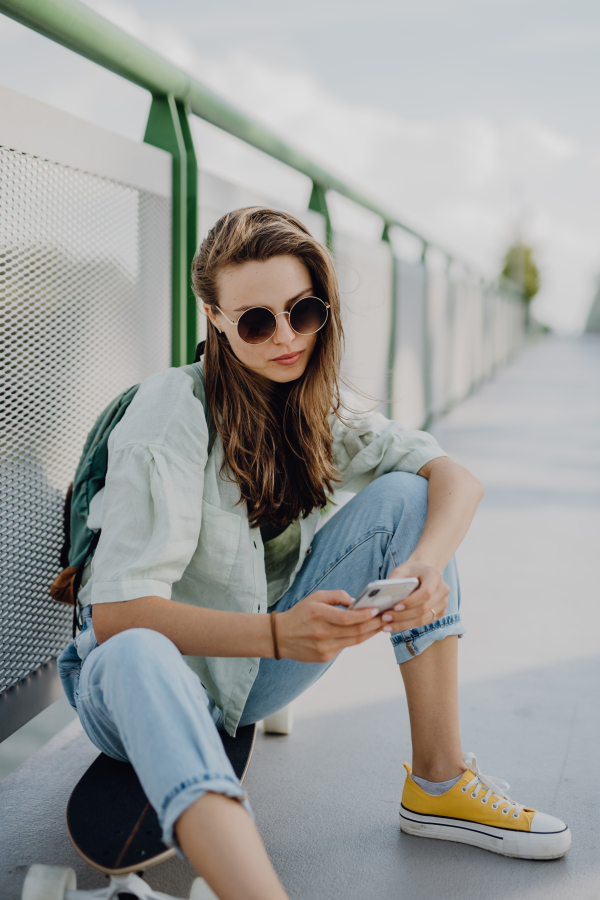 Happy young woman sitting on a skateboard and scrolling her phone at city bridge. Youth culture and commuting concept.