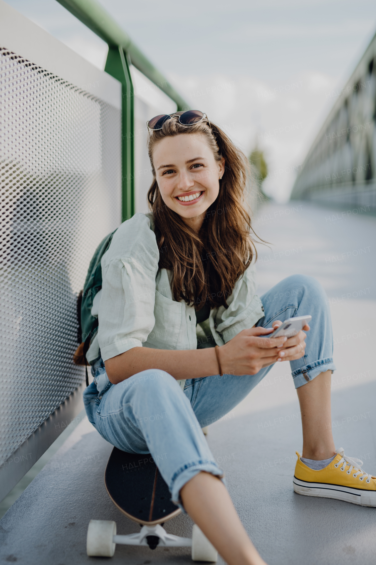 Happy young woman sitting on a skateboard at city bridge. Youth culture and commuting concept.