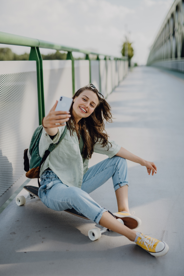 Happy young woman sitting on a skateboard and taking selfie at city bridge. Youth culture and commuting concept.