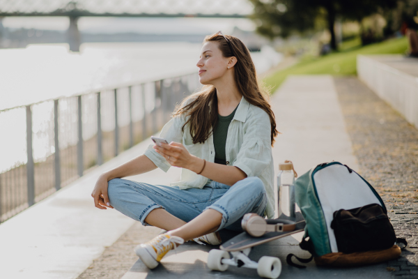 Happy young woman sitting in city park and scrolling smartphone. Youth culture and commuting concept.