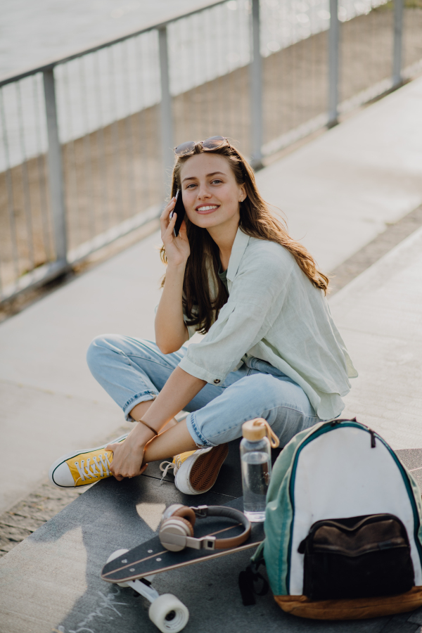 Happy young woman sitting on a skateboard and calling at city bridge. Youth culture and commuting concept.