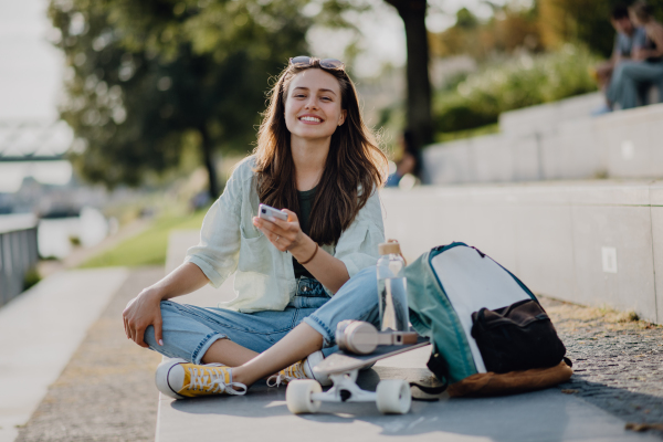 Happy young woman sitting in city park and scrolling smartphone. Youth culture and commuting concept.