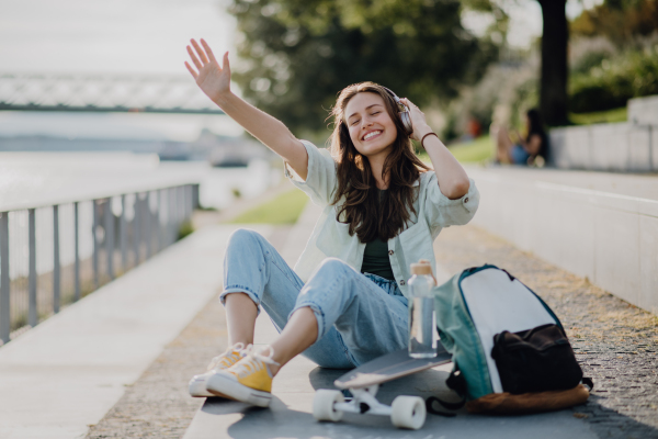 Happy young woman sitting on a skateboard and listening music at city bridge. Youth culture and commuting concept.