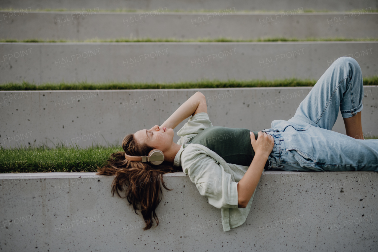 Young woman resting in city park with closed eyes, enjoying the music trough headphones.