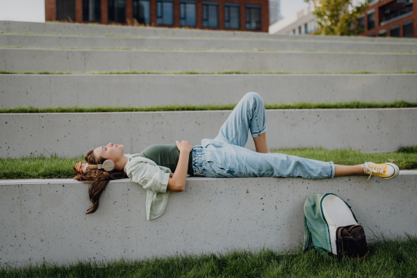 Young woman resting in city park with closed eyes, enjoying the music trough headphones.