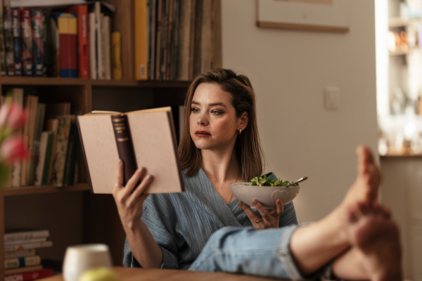 Woman resting at home, reading a book and eating salad.