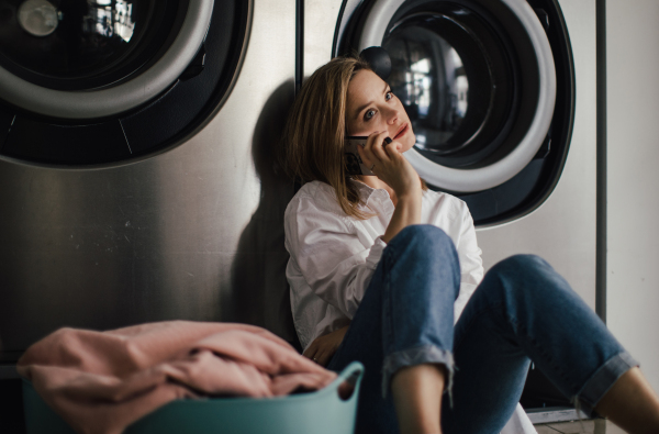 Young woman calling with smartphone, sitting in a laundry room.