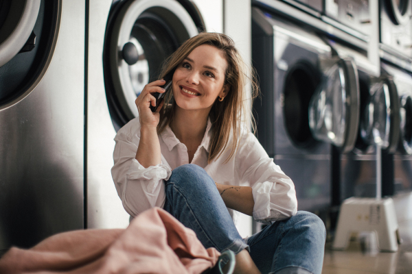 Young woman calling with smartphone, sitting in a laundry room.