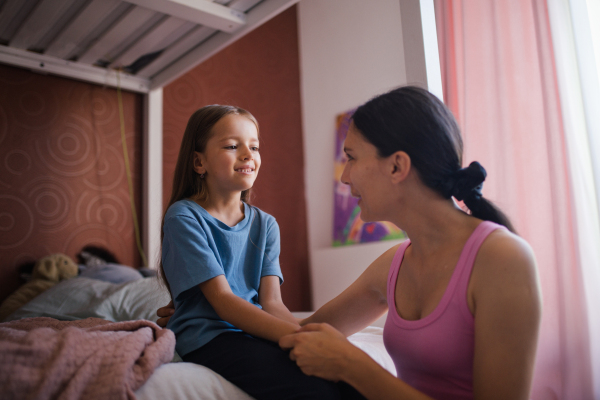 Mother waking her daughter up for school in the morning. She kneeling by the child's bed and holding her adorable daughter by hands.