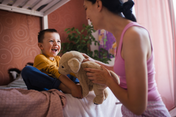 A single mother waking her son up for school in the morning. Beautiful mom awakens the schoolboy with a stuffed toy, playing and making him laugh.