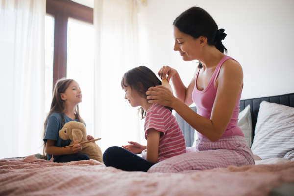 Portrait of a mother with two daughters on a bed in the bedroom. Mother combs her daughter's long hair with a comb. Concept of Mother's Day and maternal love.