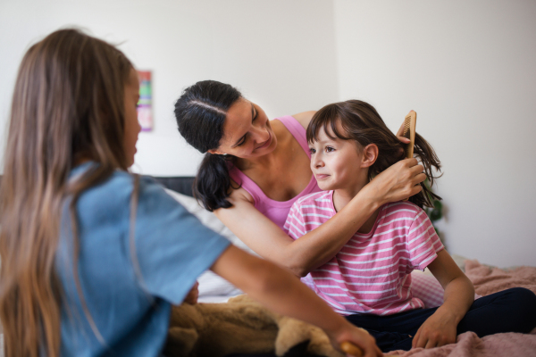 Portrait of a mother with two daughters on a bed in the bedroom. Mother combs her daughter's long hair with a comb. Concept of Mother's Day and maternal love.