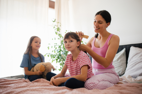 Portrait of a mother with two daughters on a bed in the bedroom. Mother combs her daughter's long hair with a comb. Concept of Mother's Day and maternal love.