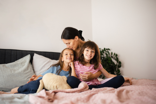 Portrait of a mother with two daughters on a bed in the bedroom. Fatherless family sit and hug each other. Concept of Mother's Day and maternal love.