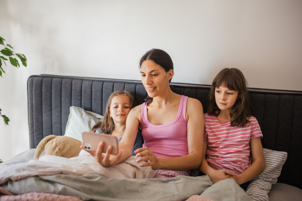 Portrait of a mother with two daughters lying on bed under blanket. Family making video call on smartphone to father. Concept of Mother's Day and maternal love.