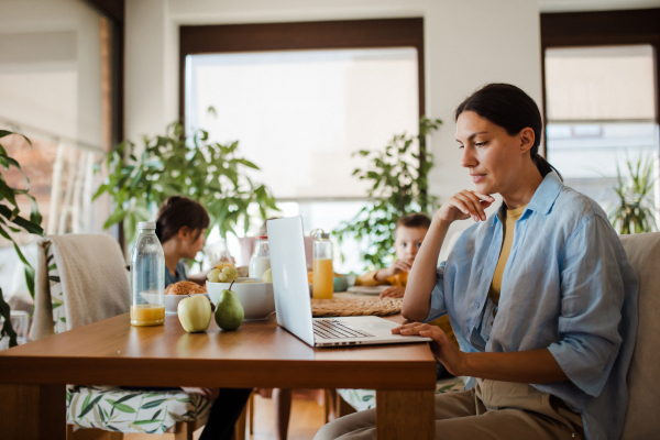 Mother working on a laptop at the kitchen table while the children have breakfast. Remote work and home office for mothers with children.