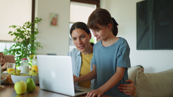 Mother working on a laptop at the kitchen table while the children have breakfast. Remote work and home office for mothers with children.