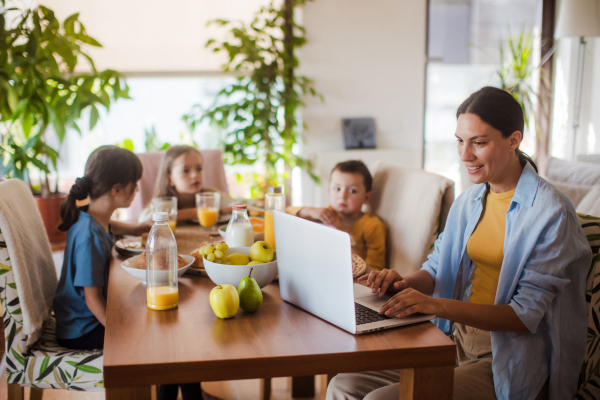 Mother working on a laptop at the kitchen table while the children have breakfast. Remote work and home office for mothers with children.