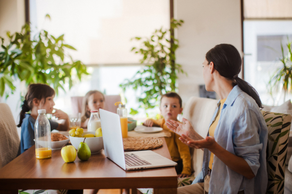 Mother working on a laptop at the kitchen table while the children have breakfast. Remote work and home office for mothers with children.