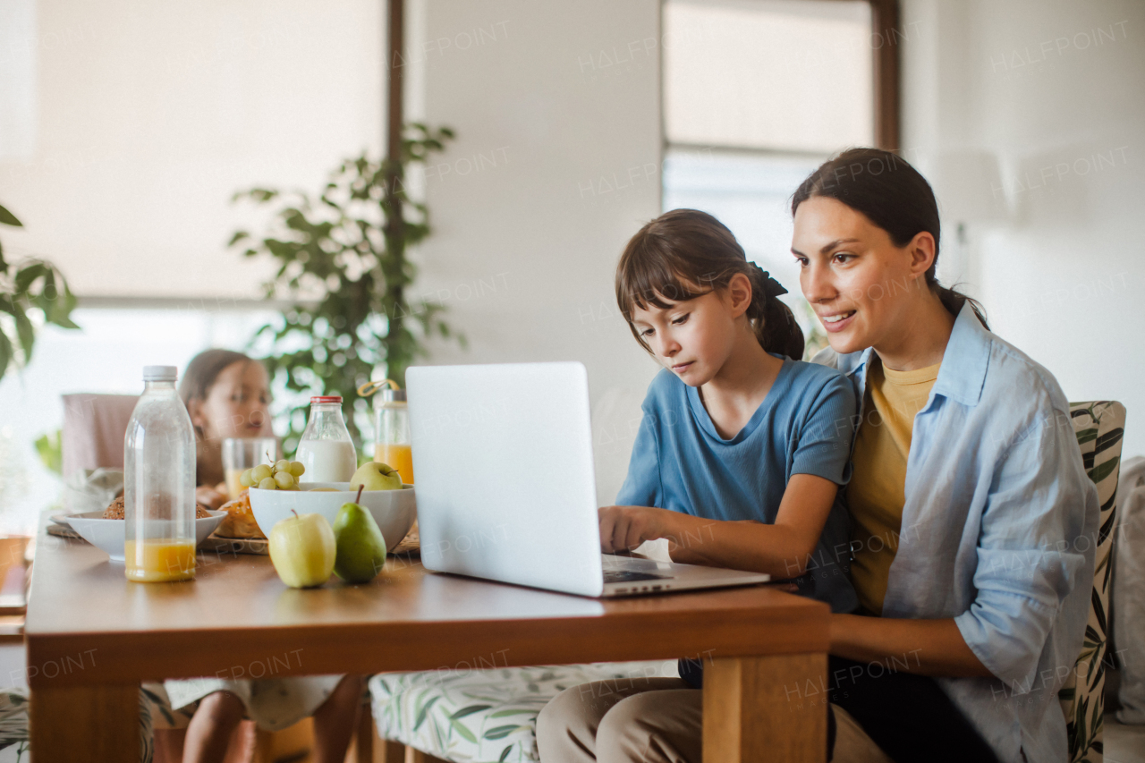 Mother working on a laptop at the kitchen table while the children have breakfast. Remote work and home office for mothers with children.