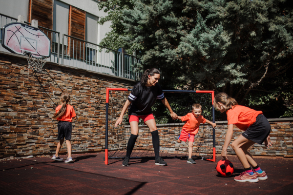 Mom playing football with her children, dressed in football jerseys. The family as one soccer team. Fun family sports activities outside in the backyard or on the street.