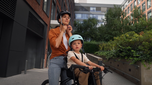 Mother carring her son on a secure child bike carrier or seat, both wearing helmets. Mom commuting with a young child through the city on a bicycle.