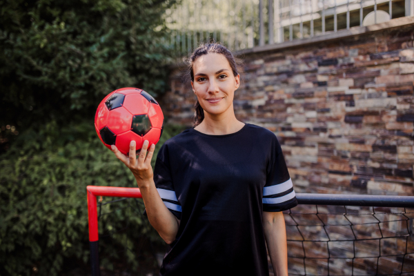 Beautiful female football player standing by the football goal with a ball in her hand. The woman plays soccer for relaxation. Concept of mental health and sport.