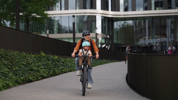 Mother carring her son on a secure child bike carrier or seat, both wearing helmets. Mom commuting with a young child through the city on a bicycle.