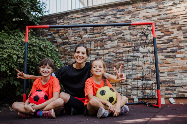 Mom playing football with her daughters, dressed in football jerseys. The family as one soccer team. Fun family sports activities outside in the backyard or on the street.