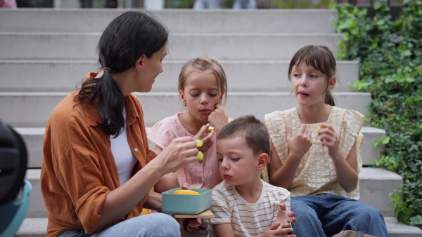 Children and mother have a healthy snack in the city after school. The family sitting on concrete steps in the city, resting and eating home made snack, fruit.