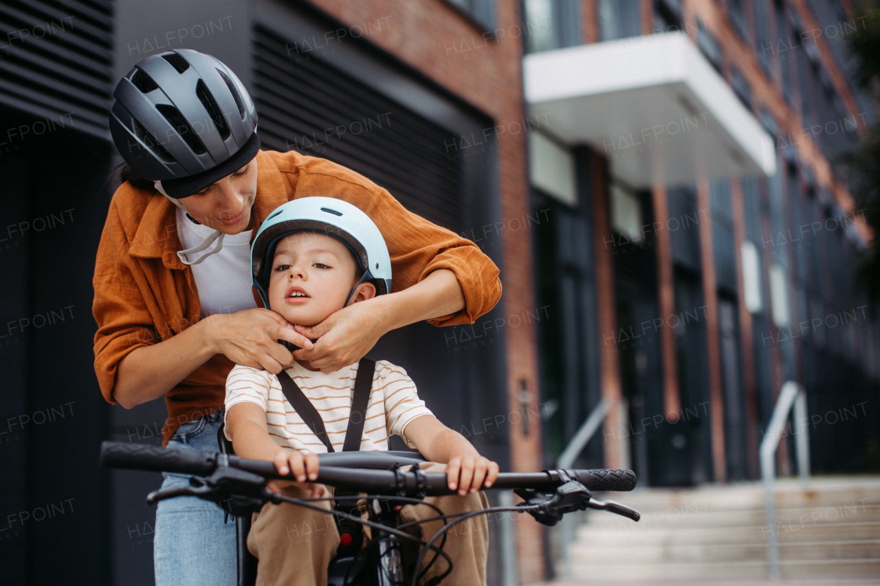 Mother fastening sons' bike helmet on head, carring him on child bike carrier or seat. Mom commuting with a young child through the city on a bicycle.