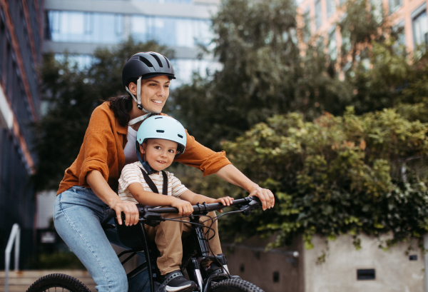 Mother carring her son on a secure child bike carrier or seat, both wearing helmets. Mom commuting with a young child through the city on a bicycle.