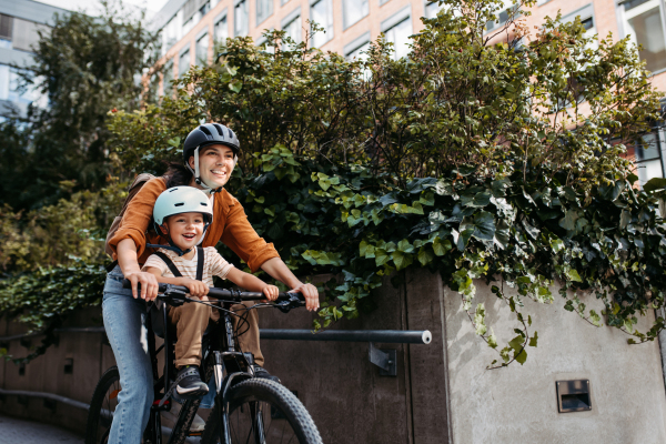 Mother carring her son on a secure child bike carrier or seat, both wearing helmets. Mom commuting with a young child through the city on a bicycle.