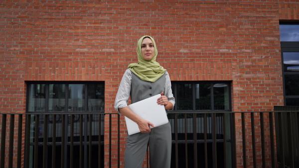 Portrait of beautiful woman in hijab standing on city street. Muslim businesswoman with laptop in hands. Iran, Afganistan female teacher in front of school building.