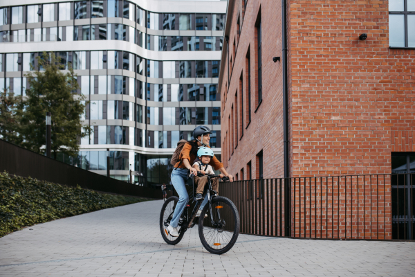 Mother carring her son on a secure child bike carrier or seat, both wearing helmets. Mom commuting with a young child through the city on a bicycle.