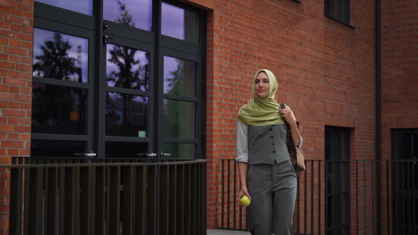 Portrait of beautiful woman in hijab standing on city street. Muslim studnet eating apple, going to school. Iran, Afganistan female teacher in front of school building.