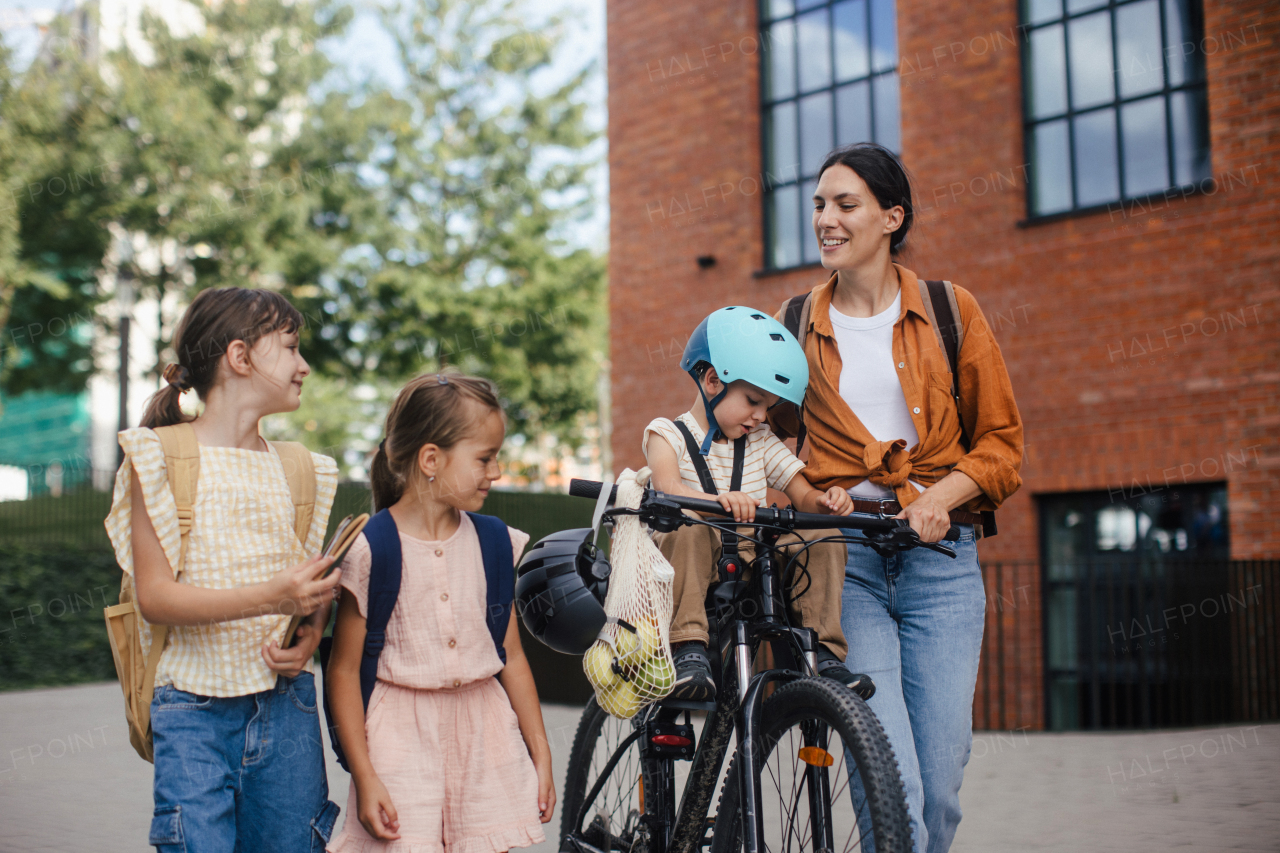 Mom picking up daughters from school with little son in bike seat . The schoolgirls telling mother about their day at school, walking in front of the school building.