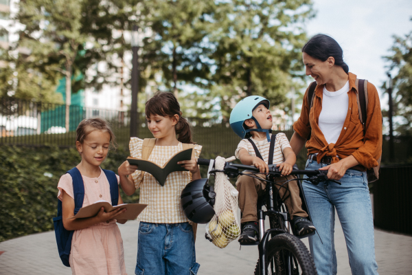 Mom picking up daughters from school with little son in bike seat. The schoolgirls telling mother about their day at school, walking in front of the school building.