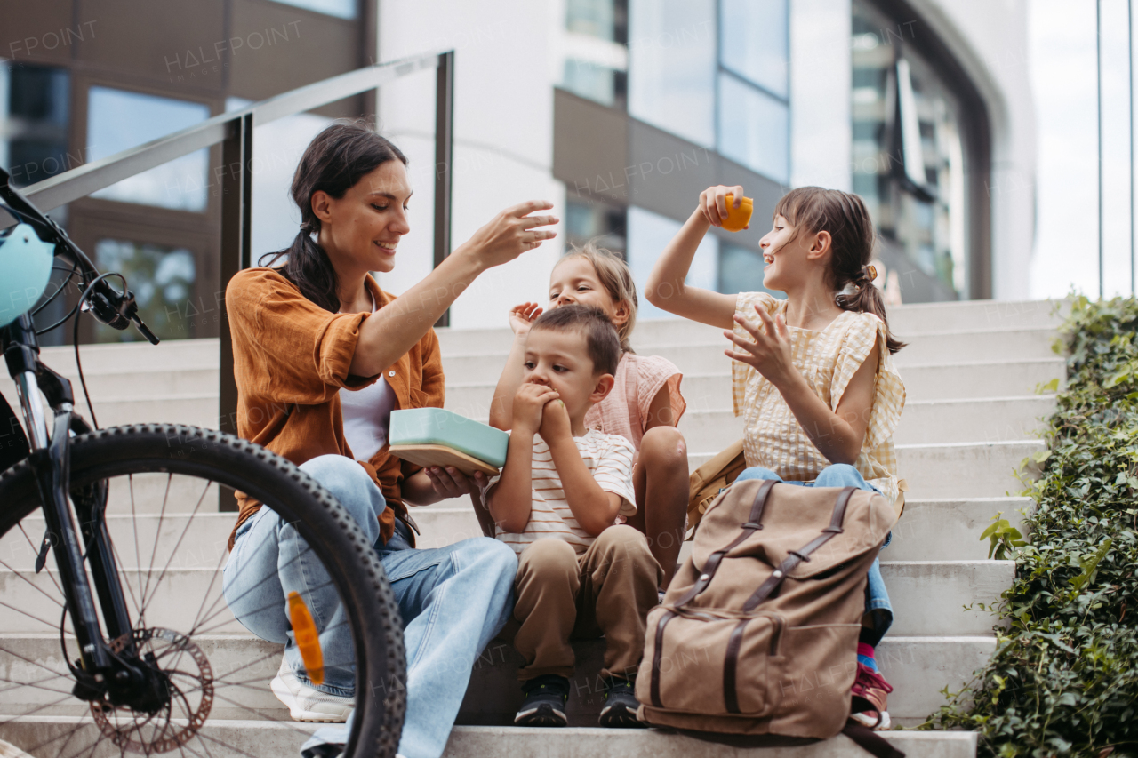 Children and mother have a healthy snack in the city after school. The family sitting on concrete steps in the city, resting and eating home made snack, fruit.