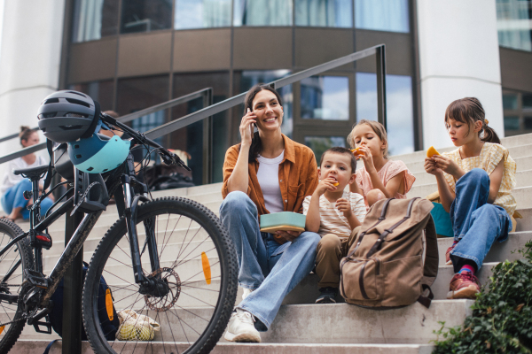 Children and mother have a healthy snack in the city after school. The family sitting on concrete steps in the city, resting and eating home made snack, while mom is on the phone.