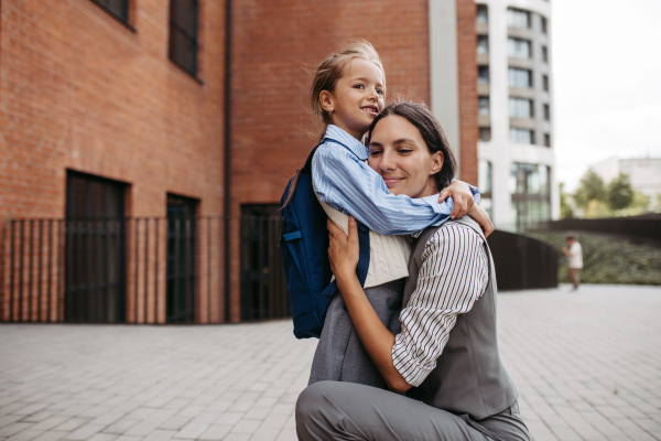 A working mother hugging daughter in front of the school building, and heading to work. Concept of work-life balance for women.